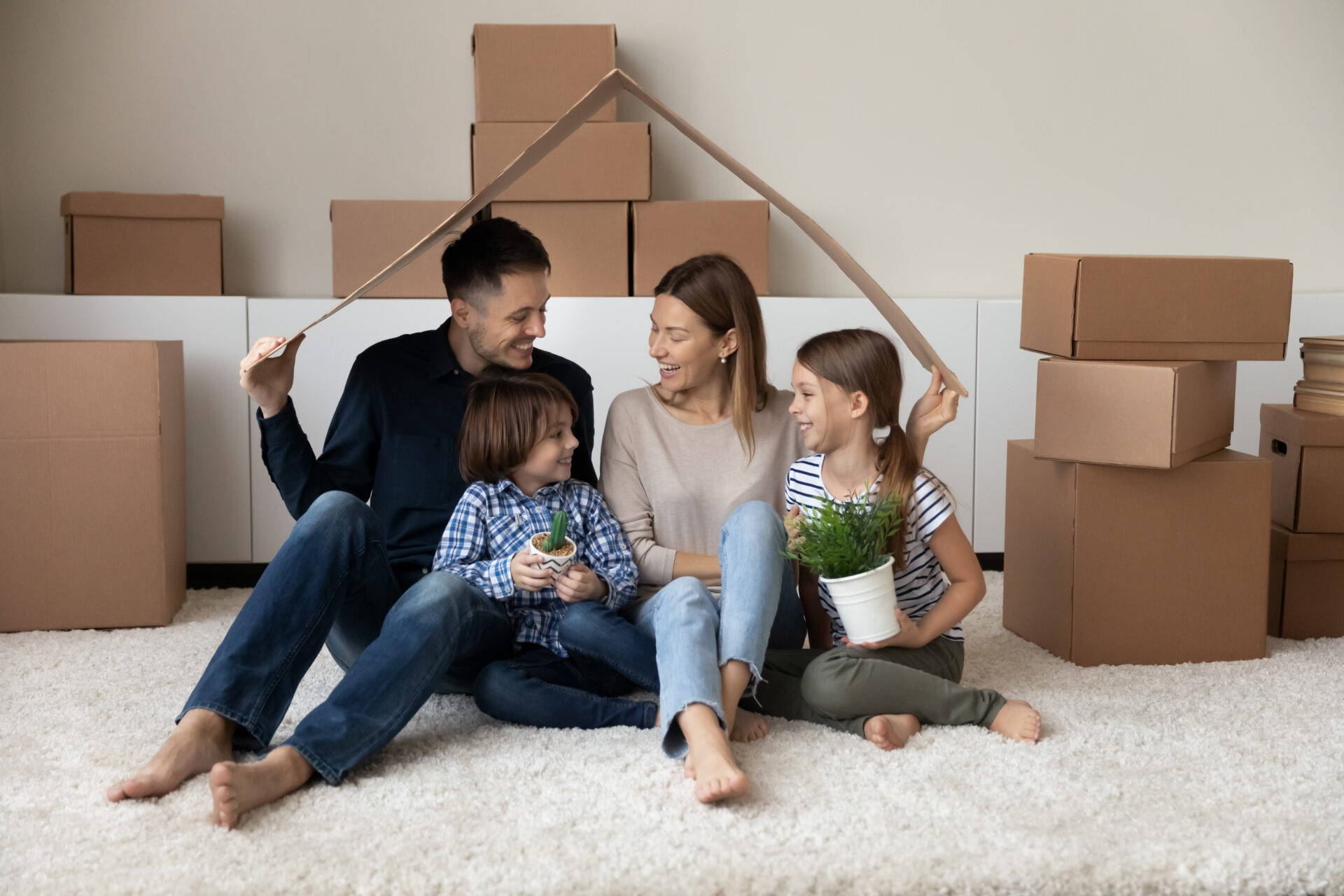 Een gezin van vier viert de verhuizing naar een nieuw huis. De ouders en twee kinderen zitten op een tapijt in een woonkamer omringd door kartonnen dozen. Ze houden een geïmproviseerd dak van een kartonnen strook boven zich, lachend van vreugde.